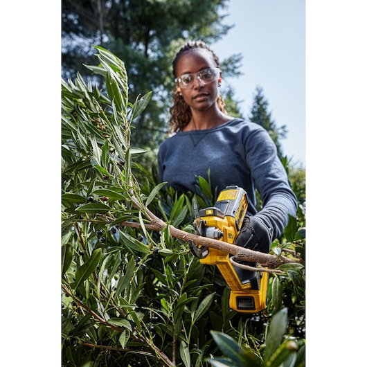 Person uses a DEWALT cordless pruner to cut a tree branch outdoors, surrounded by green foliage.