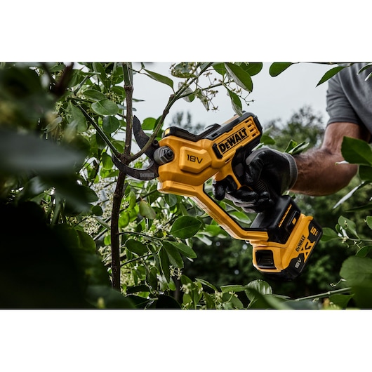 DEWALT 18V cordless pruner in use, cutting a tree branch outdoors with a gloved hand.