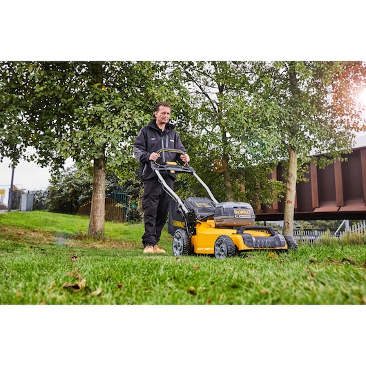 Person using a DEWALT DCMW564P2 lawn mower outdoors on grass near trees.