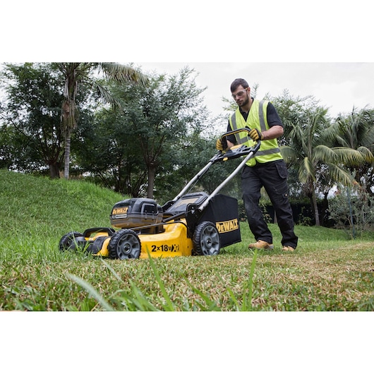 Person mowing grass with a DEWALT cordless lawn mower outdoors. Trees and greenery in background.