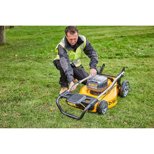 Person adjusts a DEWALT DCMW564P2 cordless lawn mower on grass, wearing a safety vest and work boots.