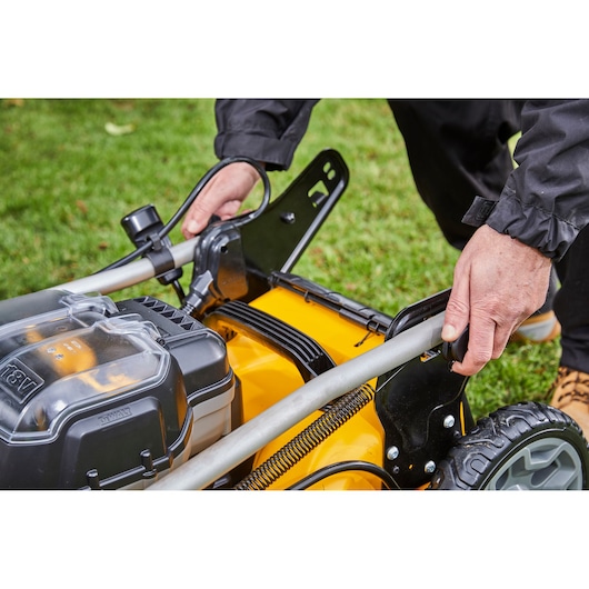 Close-up of hands adjusting the handle on a DEWALT cordless lawn mower outdoors on grass.