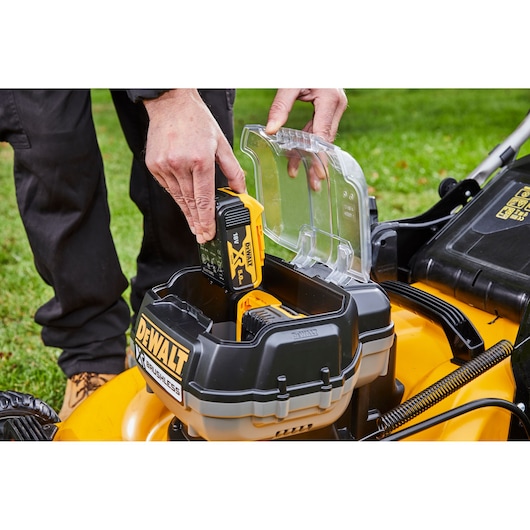 Hand inserting DEWALT battery into a lawn mower's battery compartment, outdoors on grass.
