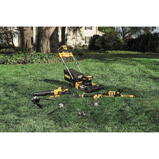 DEWALT cordless lawn mower and garden tools are displayed on grass in a yard outside a house.