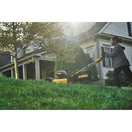 Person mowing lawn with a DEWALT cordless mower near a house on a sunny day.