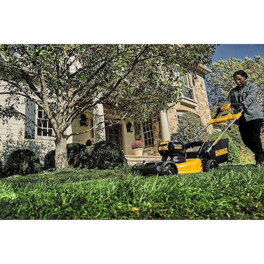 Person using a DEWALT lawn mower on grass in front of a house with trees and bushes on a sunny day.