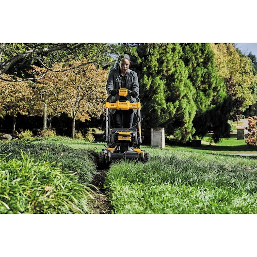 Person using a DEWALT lawn mower outdoors, cutting grass in a green garden with trees in the background.