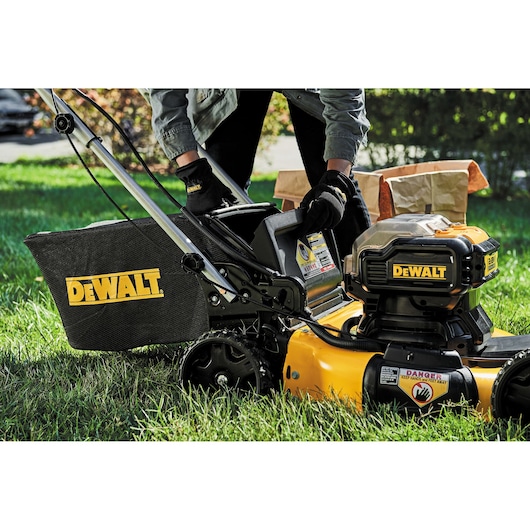 Person inserts battery into yellow DEWALT lawn mower on grass, with lawn bags in the background.