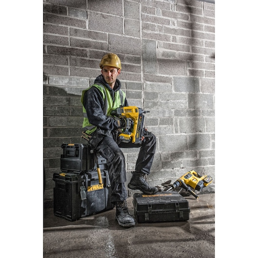 Worker with DEWALT DCN890P2 nailer sitting on toolboxes at a construction site with safety gear.