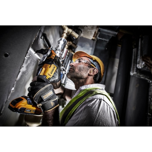 Person using a DEWALT DCS355D2 cordless tool to cut metal piping in an industrial setting, wearing safety gear.