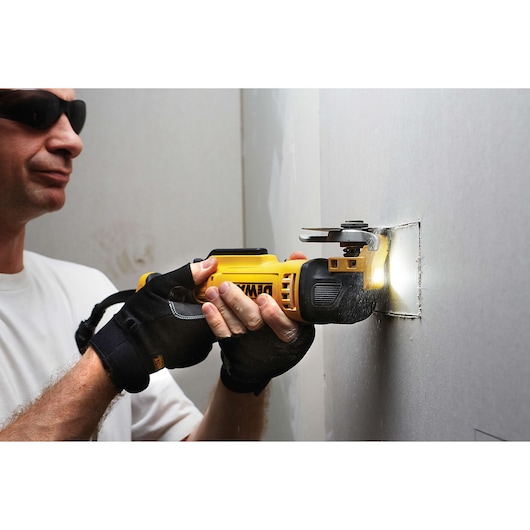 Gloved hands use a DEWALT oscillating tool to cut a square hole in a wall, tool emits light at the cut.