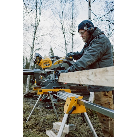 Person using DEWALT DHS780T2 saw outdoors to cut wooden plank, wearing safety gear and gloves.