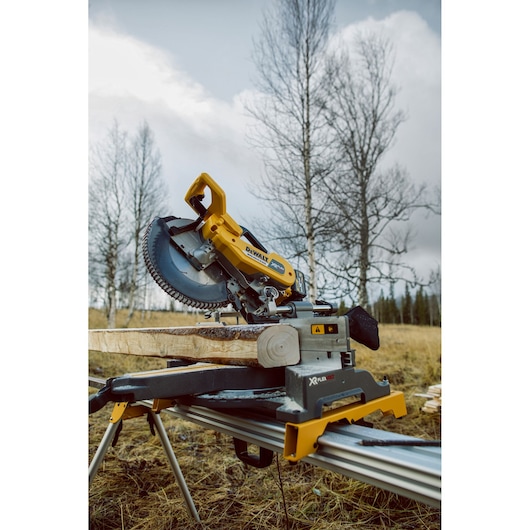 DEWALT miter saw cutting wood outdoors on a stand, with trees and cloudy sky in the background.