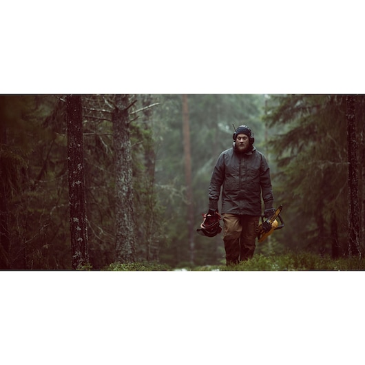 Person walking in a forest, carrying a DEWALT saw and safety gear, surrounded by trees and greenery.