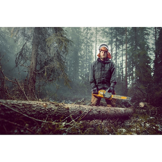 Person in safety gear holding DEWALT chainsaw in forest, standing by a fallen tree trunk.