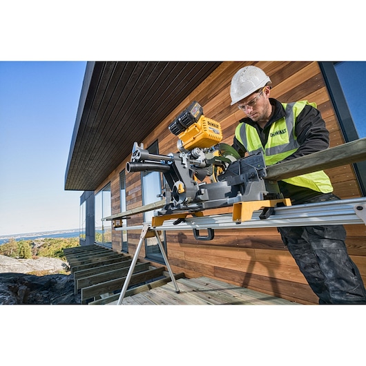 Worker uses DEWALT DHS780T2 saw outdoors on wooden deck at a construction site, wearing safety gear.