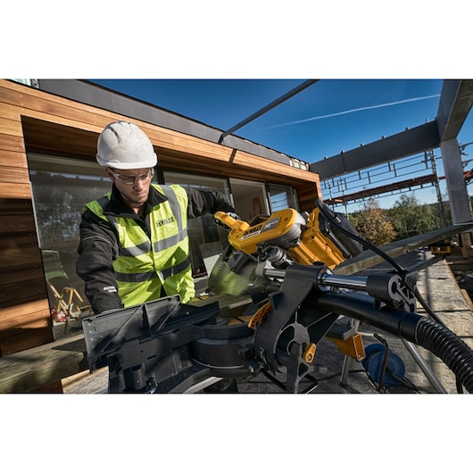 Person using a DEWALT miter saw outdoors at a construction site, wearing safety gear and a high-visibility vest.