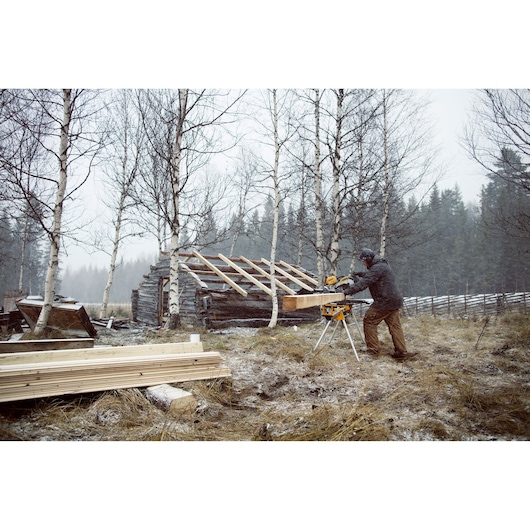 Person uses DEWALT saw outdoors in winter near a cabin under construction, surrounded by trees and lumber.