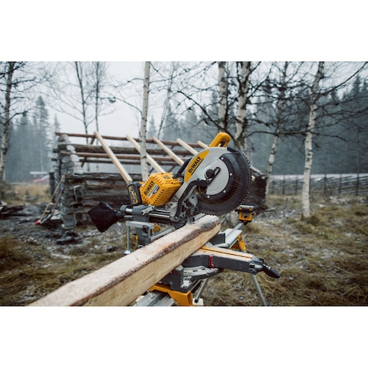 DEWALT miter saw cutting wood outdoors near a rustic building site with trees in the background.