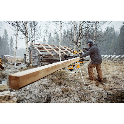 Person using a DEWALT saw to cut a long wooden beam outdoors near a log cabin on a snowy day.