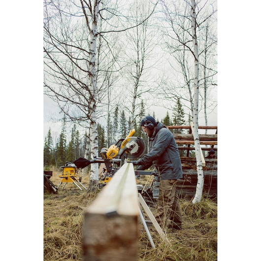 Person cutting wood outdoors with a DEWALT miter saw, surrounded by birch trees and forest background.