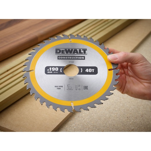 Hand holding DEWALT DT1945 190mm 40T circular saw blade for construction with wood in background.