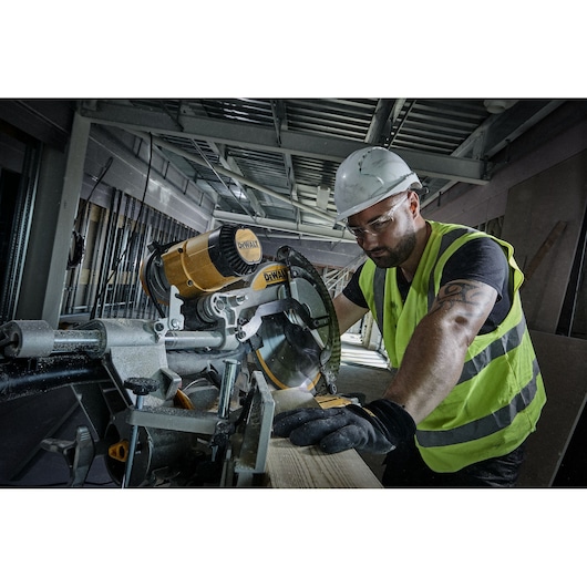 Worker using a DEWALT circular saw to cut wood at a construction site, wearing safety gear and high-visibility vest.