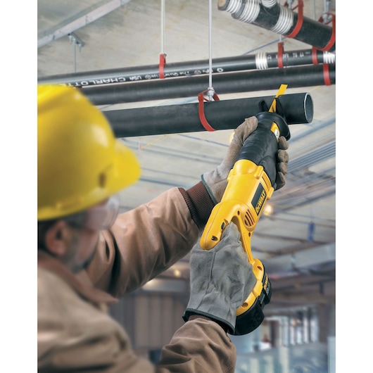 Worker uses a DEWALT reciprocating saw to cut a black pipe overhead in an industrial setting.