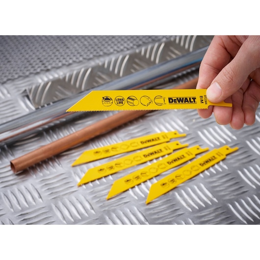 A hand holds a yellow DEWALT DT2384 saw blade with others lying on metal sheet beside metal pipes.