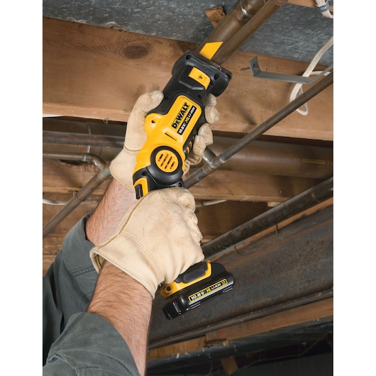 Gloved hands use a DEWALT cordless reciprocating saw to cut a pipe beneath wooden beams.
