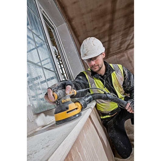 Worker using DEWALT DT3101 sander on a windowsill at a construction site, wearing safety gear and helmet.