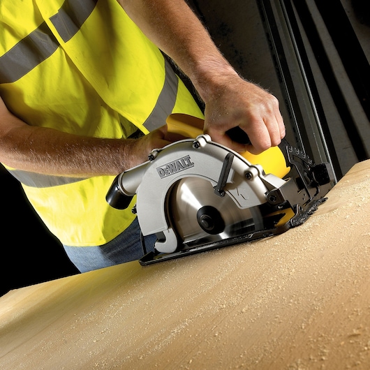 Person using a DEWALT circular saw to cut wood. Sawdust covers the surface. High-vis vest is visible.