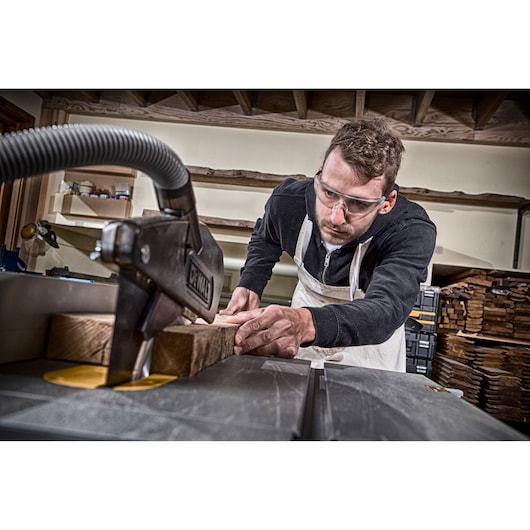 Person uses a DEWALT table saw to cut wood in a workshop, wearing a white apron. Wood is stacked in background.