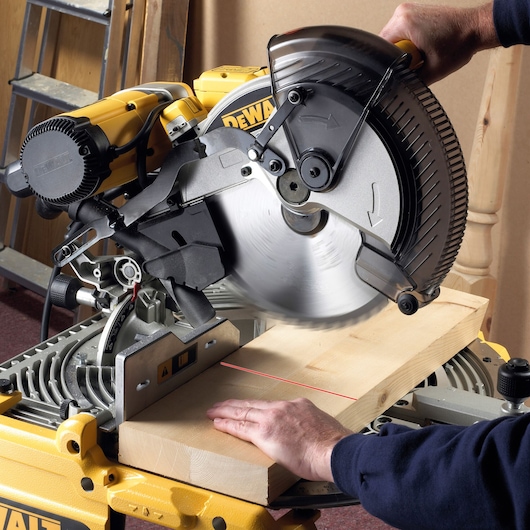 A DEWALT miter saw being used to cut a wooden plank. The saw blade is spinning and a person's hand is holding the plank steady on the saw table. The saw has a yellow and black design typical of DEWALT tools.