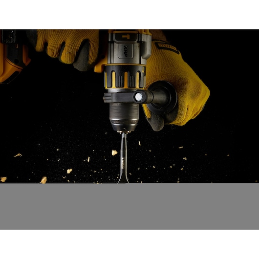 Gloved hands using a DEWALT drill and spade bit to drill into wood with scattered wood chips.