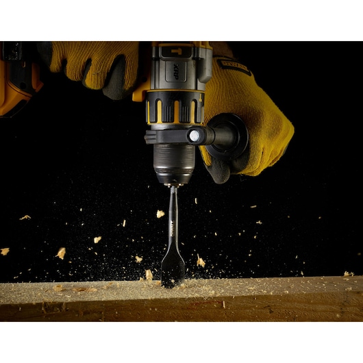 Close-up of a person wearing yellow gloves using a DEWALT power drill with a flat wood drill bit to drill into a wooden surface, with wood chips flying.