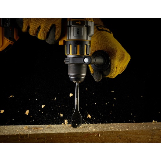 Gloved hands using a DEWALT drill with a spade bit to drill into wood, wood chips flying.