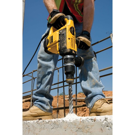 Worker uses a DEWALT drill to bore into concrete at a construction site, wearing gloves, jeans, and boots.