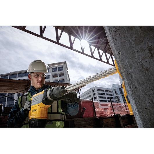 Worker uses DEWALT drill with DT60811 bit on concrete at urban construction site; modern buildings in background.