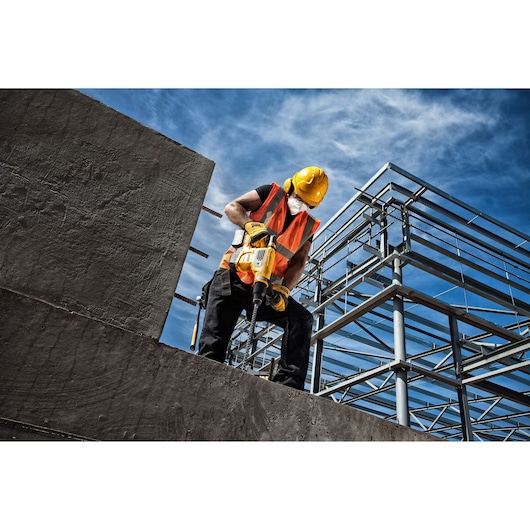 Worker in safety gear uses yellow tool on concrete at construction site with steel framework under blue sky.