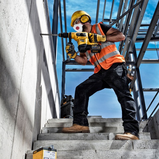 Worker uses DEWALT drill on concrete wall. SKUs: DT60814, DT60814-QZ. Construction site, safety gear visible.
