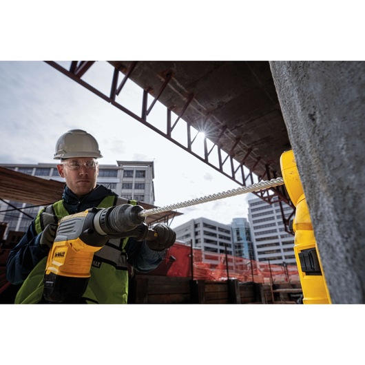 Worker uses a DEWALT drill with a long bit at a construction site with buildings in the background.