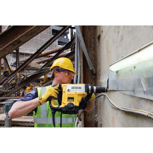 Worker uses DEWALT rotary hammer drill to drill into a wall at a construction site. Safety gear and gloves are worn.