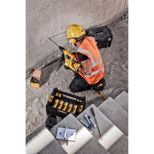 Worker in safety gear uses a DEWALT drill near concrete stairs, with cases, bits, and fasteners nearby.