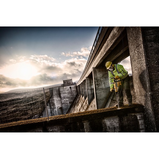 Worker using a DEWALT drill on concrete structure outdoors near a dam at sunset.
