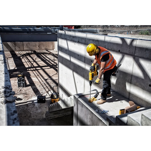 Worker uses a yellow DEWALT drill on concrete at a construction site, wearing safety gear and an orange vest.