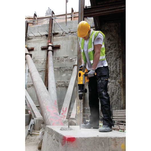 Worker uses a DEWALT drill on concrete at a construction site, wearing safety gear and a yellow helmet.
