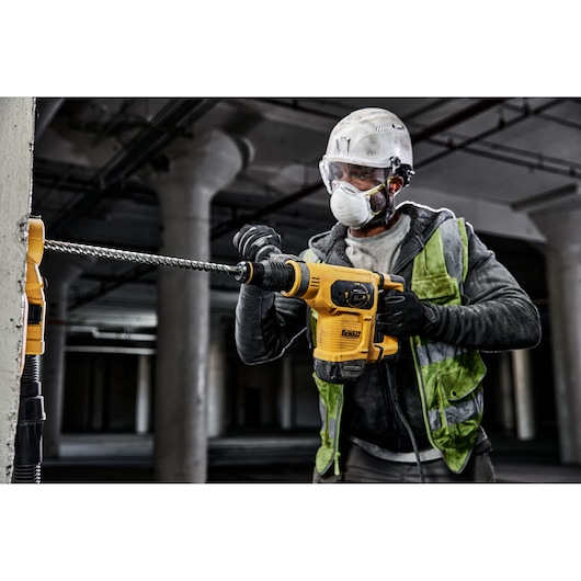 Worker drills concrete wall with DEWALT rotary hammer. Wearing safety gear and reflective vest at construction site.
