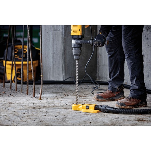 Person using DEWALT hammer drill with dust extractor on a concrete floor at a construction site.