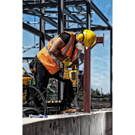 Worker in safety gear uses a DEWALT drill at a construction site with steel framework and concrete surface.